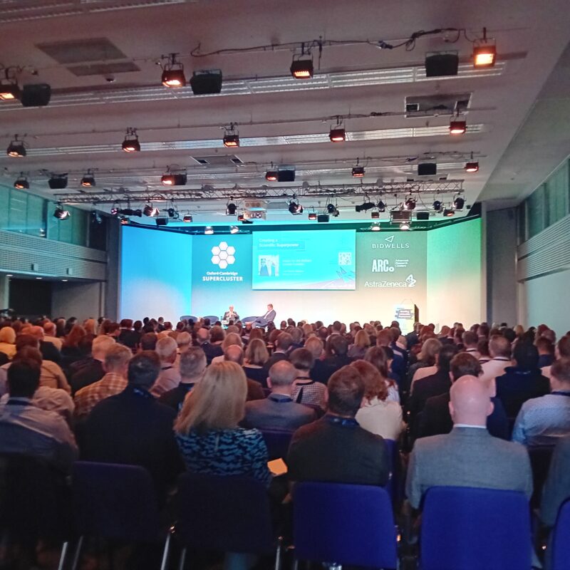 a picture from the back of a seated crowd looking toward the stage of the conference/ Two speakers are seated on stage and there is a blue screen as the background.