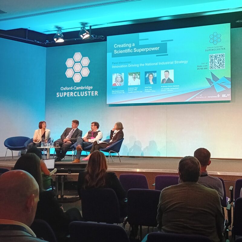 a picture from the back of a seated crowd looking toward the stage of the conference. Four speakers are seated on stage and there is a blue screen as the background.